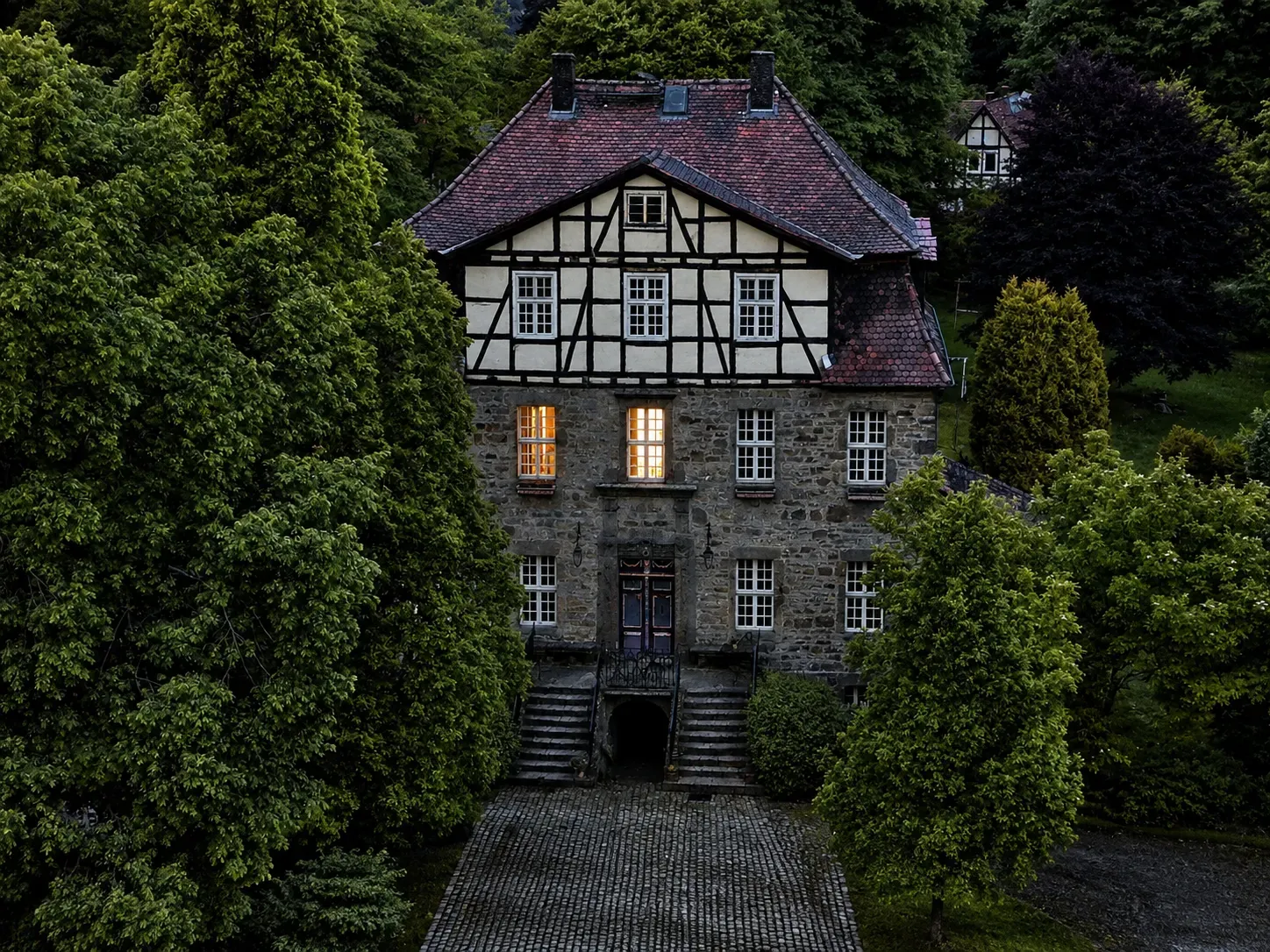 Frontansicht des Fachwerk-Gutshauses mit beleuchtetem Fenster, gerahmt von hohen Nadelbäumen